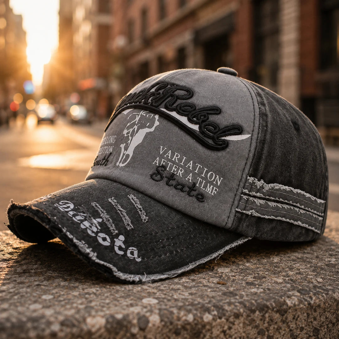Josh & Tate - BlackRebel™ Distressed Cap - A distressed gray baseball cap with black and white embroidered patches, including the word 'Rebel' and 'VARIATION AFTER A TIME State', resting on a stone surface with a blurred city street in the background.