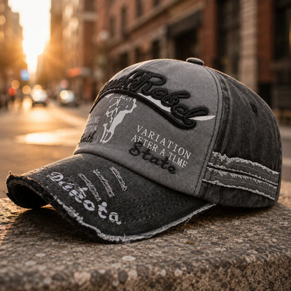 Josh & Tate - BlackRebel™ Distressed Cap - A distressed gray baseball cap with black and white embroidered patches, including the word 'Rebel' and 'VARIATION AFTER A TIME State', resting on a stone surface with a blurred city street in the background.