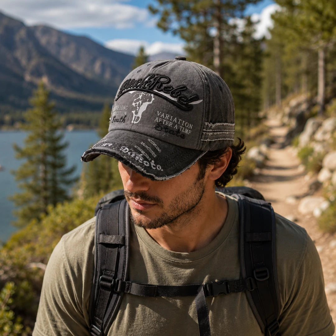 Josh & Tate - BlackRebel™ Distressed Cap - A man hiking outdoors on a trail near a lake and mountains, wearing a dark gray distressed baseball cap with embroidered text and graphics, an olive green t-shirt, and a black backpack.