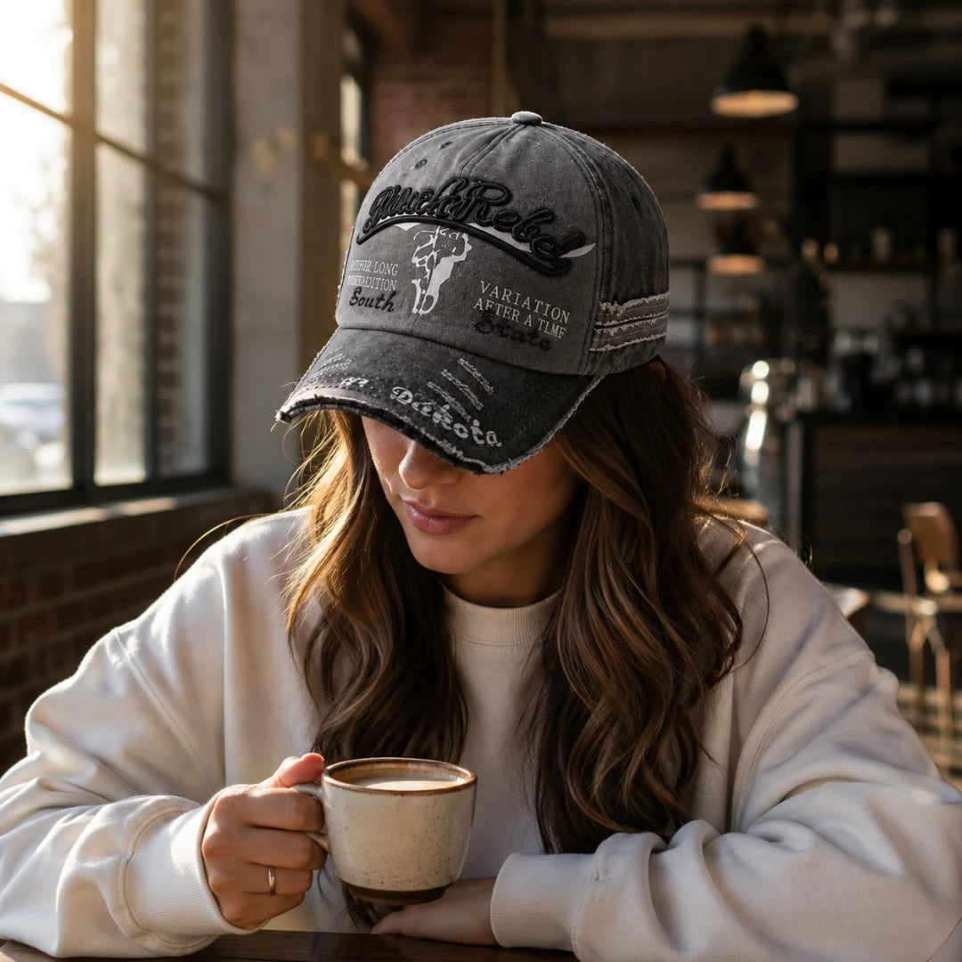 Josh & Tate - BlackRebel™ Distressed Cap - A woman in a white sweatshirt wearing a distressed dark gray baseball cap with embroidered text, sitting in a cafe and holding a mug.