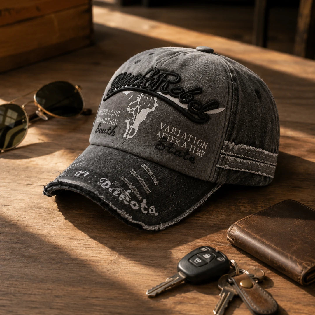 Josh & Tate - BlackRebel™ Distressed Cap - A distressed dark gray baseball cap with embroidered 'Wild Rebel' lettering and western-style graphics, styled on a wooden table with sunglasses, keys, and a wallet nearby.