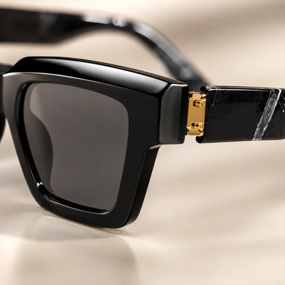 Josh & Tate - BlockFrame™ Bold Square Sunglasses - Close-up of black square sunglasses with dark lenses and a gold-tone hinge detail, shown on a warm beige background.