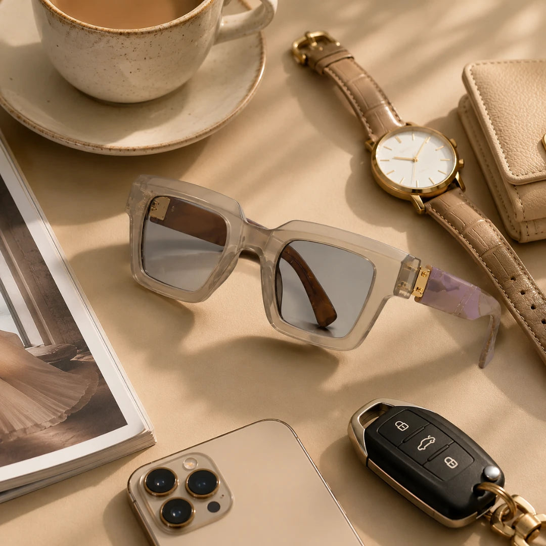 Josh & Tate - BlockFrame™ Bold Square Sunglasses - A styled flat-lay featuring a pair of translucent square sunglasses with mirrored lenses and a purple patterned temple, arranged on a beige surface alongside a watch, wallet, phone, keys, and a cup.