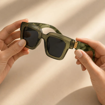 Josh & Tate - BlockFrame™ Bold Square Sunglasses - A pair of oversized square sunglasses with translucent olive green frames and dark lenses, held by two hands against a warm beige background with soft shadows.
