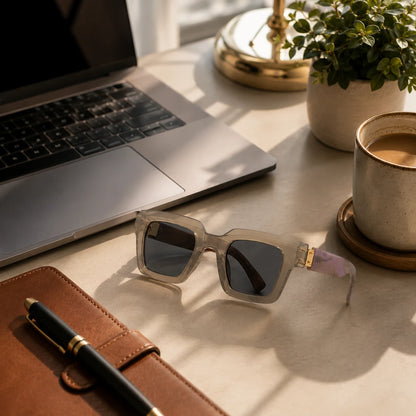 Josh & Tate - BlockFrame™ Bold Square Sunglasses - A pair of translucent light gray square sunglasses with dark lenses rests on a beige desk alongside a brown leather notebook, a black pen, a silver laptop, a potted plant, and a cup of coffee.