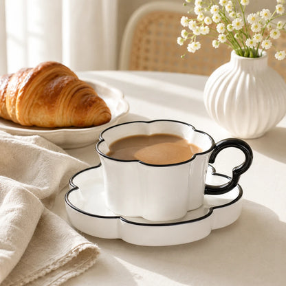 Josh & Tate - BloomCup™ Porcelain Cup & Saucer - A white ceramic coffee cup and saucer set with black scalloped edges, filled with coffee on a table. A croissant sits on a plate in the background, and a white vase with small white flowers is visible behind it.
