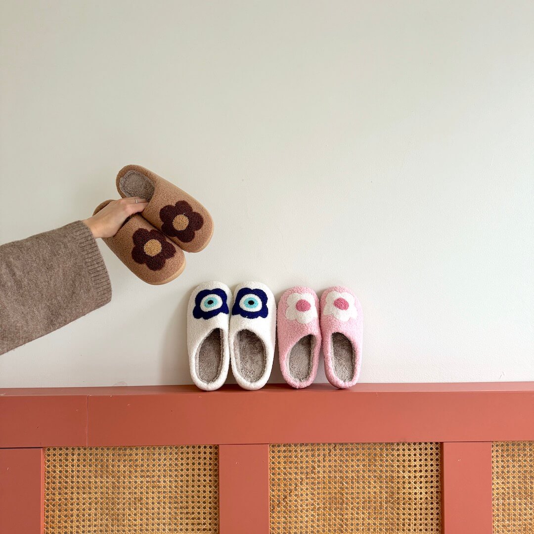 Pair of brown slippers with flower design on a white surface, with three pairs of colorful slippers below on a pink shelf.