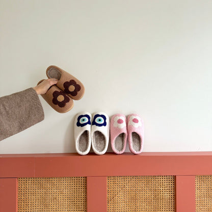 Pair of brown slippers with flower design on a white surface, with three pairs of colorful slippers below on a pink shelf.