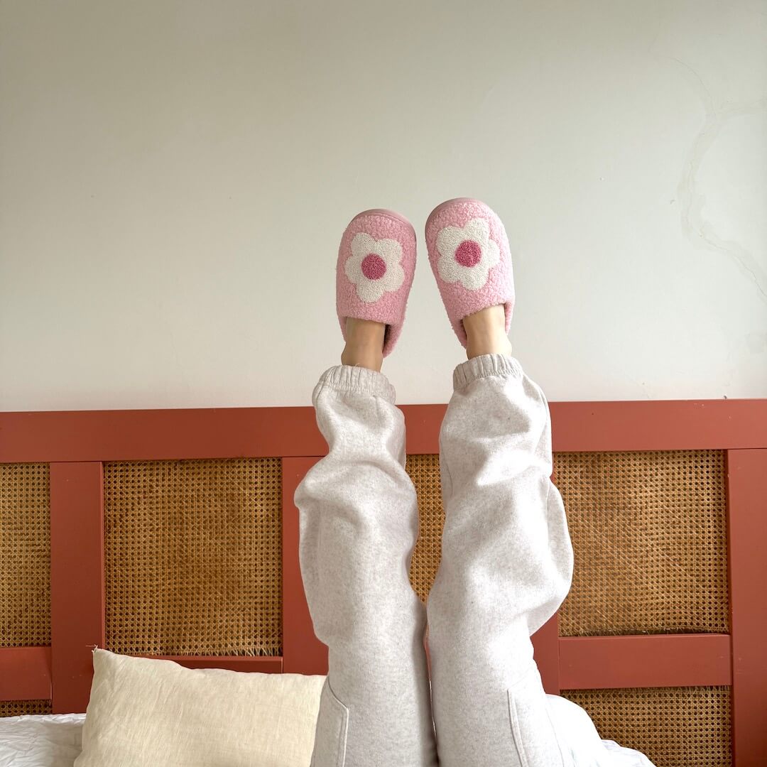 Person wearing pink slippers with floral designs and white pants, sitting on a bed.