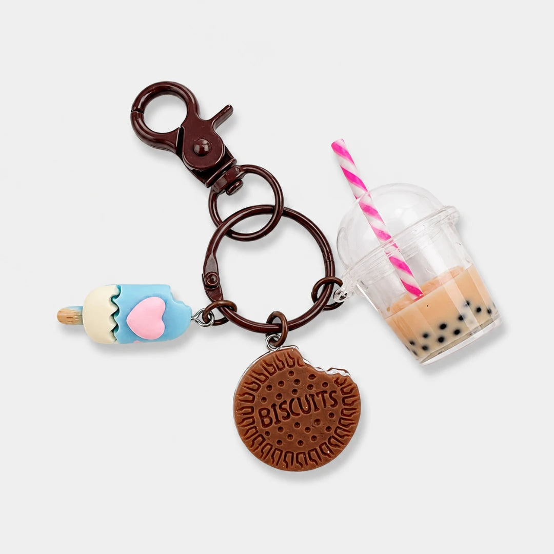 Josh & Tate - BubbleBite™ Milk Tea Keychain - A novelty keychain featuring a brown plastic clasp, a round brown biscuit charm engraved with 'BISCUITS', a light blue popsicle charm with a pink heart, and a clear cup charm filled with bubble tea and a pink-and-white striped straw, all on a light gray background.