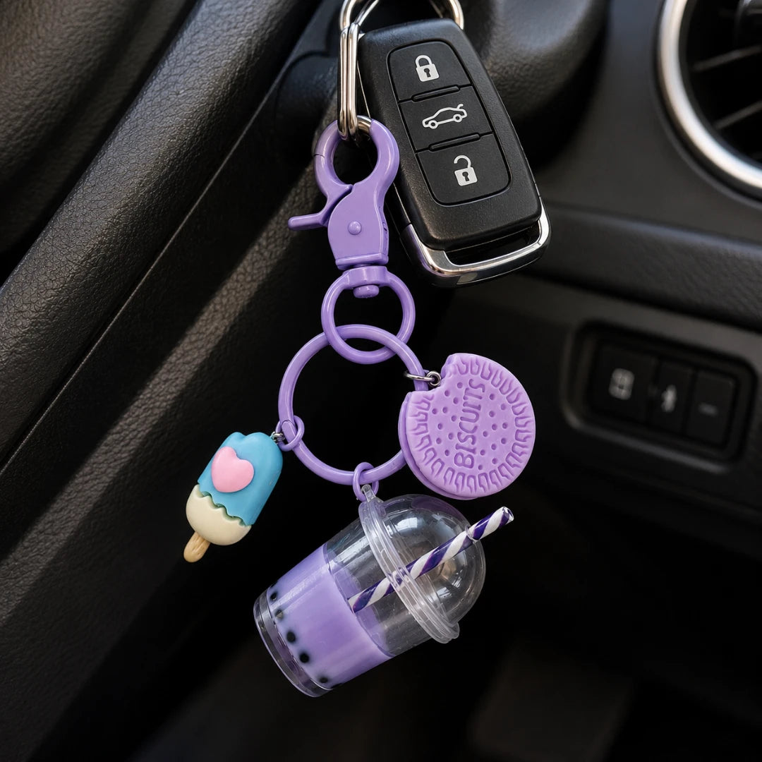 Josh & Tate - BubbleBite™ Milk Tea Keychain - A purple silicone keychain accessory set hanging from a car key fob, featuring a clasp, key rings, a purple cookie charm, a blue popsicle charm, and a small purple drink cup charm, shown in a car interior.