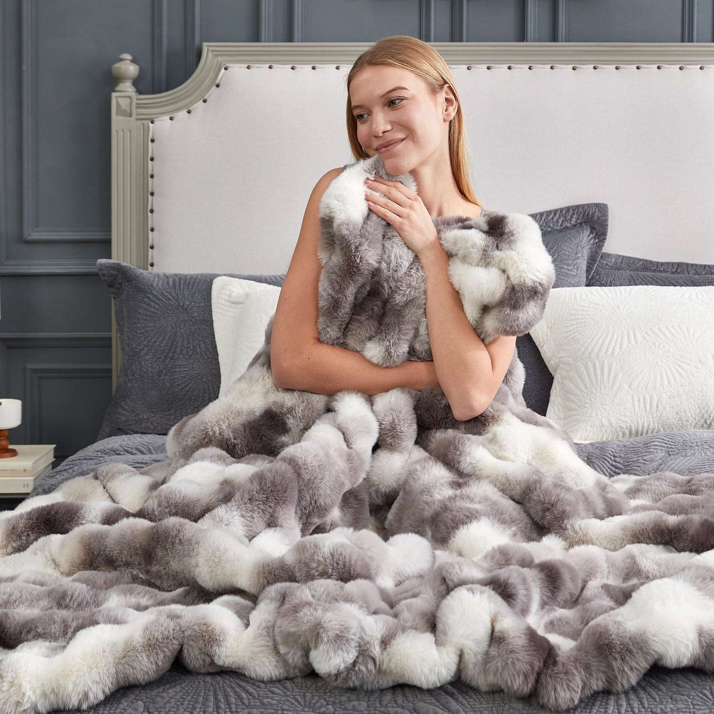 Woman wrapped in a fluffy gray blanket on a bed with a gray headboard.