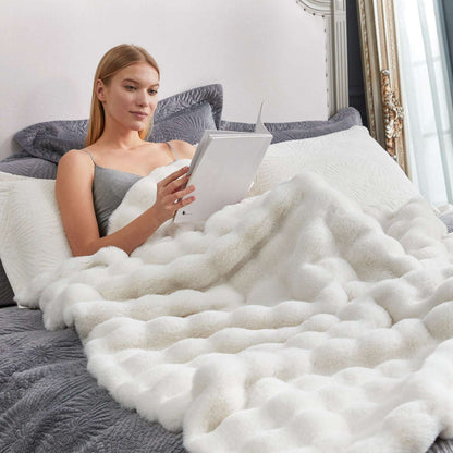 Woman reading a book under a fluffy white blanket on a bed.