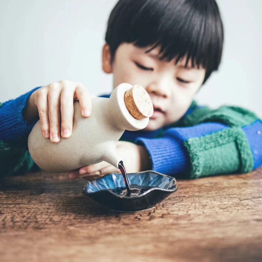 Josh & Tate - Ceramic Condiment Storage Set - Child pouring from a ceramic bottle with a cork stopper into a small dish on a wooden surface.