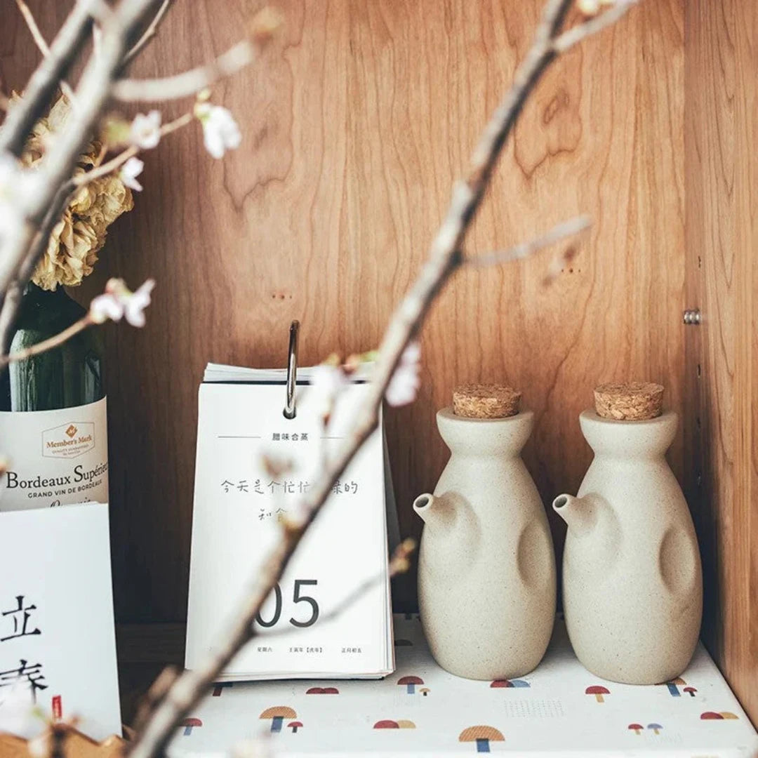 Josh & Tate - Ceramic Condiment Storage Set - Two ceramic bottles with cork stoppers on a wooden shelf with a calendar and wine bottle in the background.