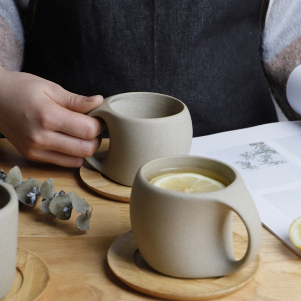 Person holding a ceramic mug with another mug and lemon on a wooden table.