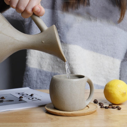 Person pouring water from a ceramic pitcher into a matching mug on a wooden table with a lemon and magazine in the background.