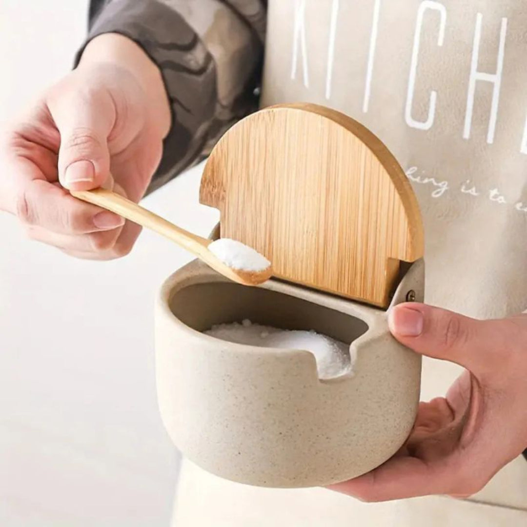 Person using a wooden spoon to scoop salt from a ceramic container with a wooden lid.