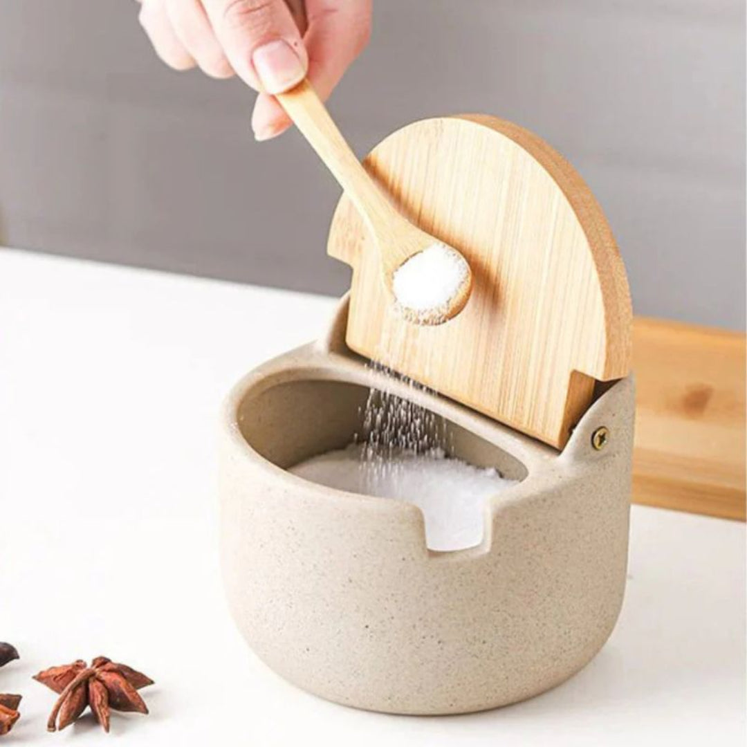 Person using a wooden spoon to scoop salt into a ceramic container on a neutral background