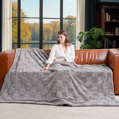 Woman sitting on a brown leather couch with a gray quilt draped over her, in a room with large windows showing trees outside.