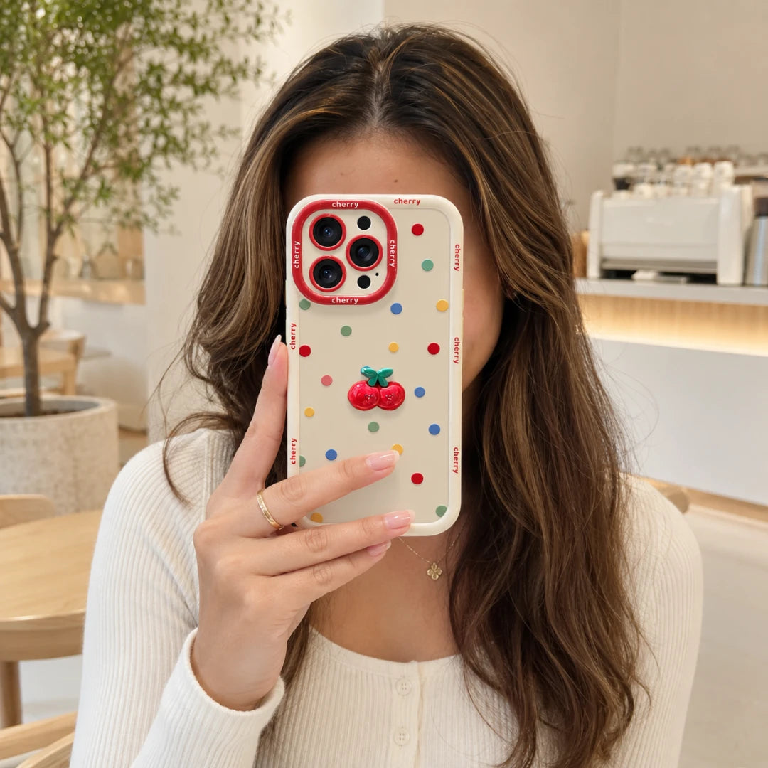 Josh & Tate - CherryPop™ iPhone Case - A woman takes a mirror selfie showing a phone case with a white base, multicolor polka dots, a red cherry 3D design, and red camera bumper rings, set against a bright cafe background.