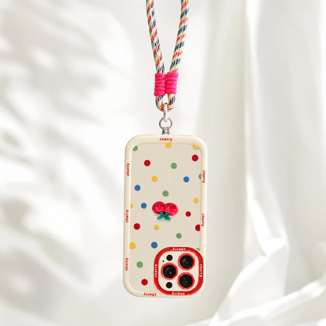 Josh & Tate - CherryPop™ iPhone Case - A clear phone case with colorful polka dots, a red cherry design, and 'Cherry' printed repeatedly along the border, hanging from a pink and multicolor braided lanyard against a soft white draped background.