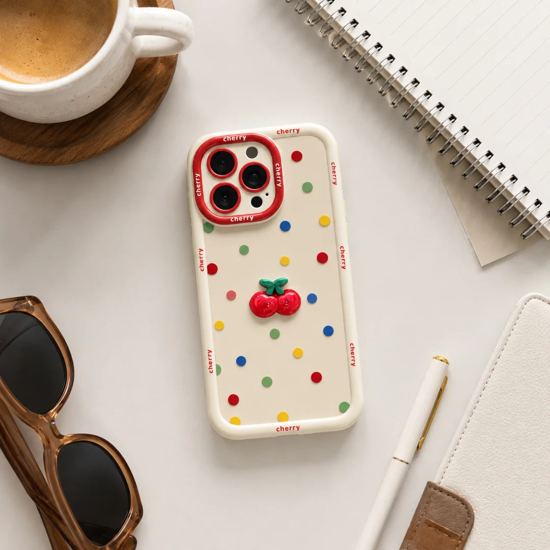 Josh & Tate - CherryPop™ iPhone Case - A phone case with a white base, colorful polka dots, and a 3D cherry design in the center, shown in a styled flat-lay with a coffee cup, sunglasses, notebook, and pen.