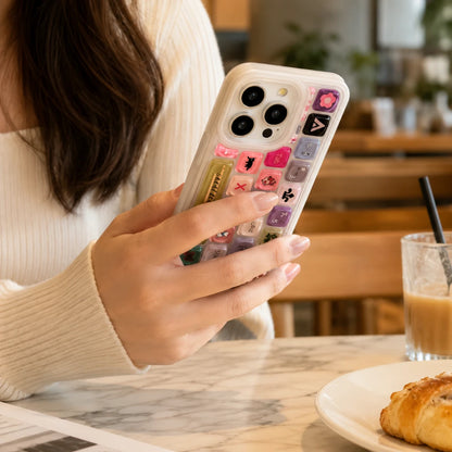 Josh & Tate - 
ClickPod™ Keycap Phone Case
  - A person in a cream ribbed sweater holds a white smartphone with a decorative case featuring colorful keyboard-style key graphics and small character motifs, photographed in a cafe setting with a drink and pastry on the table.