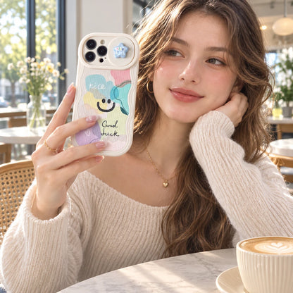 Josh & Tate - CloudPop™ Sliding Lens iPhone Case - A woman in a cozy cafe setting holds up a white phone case decorated with pastel rainbow clouds, a smiling face, and the text 'have peace Good luck'.