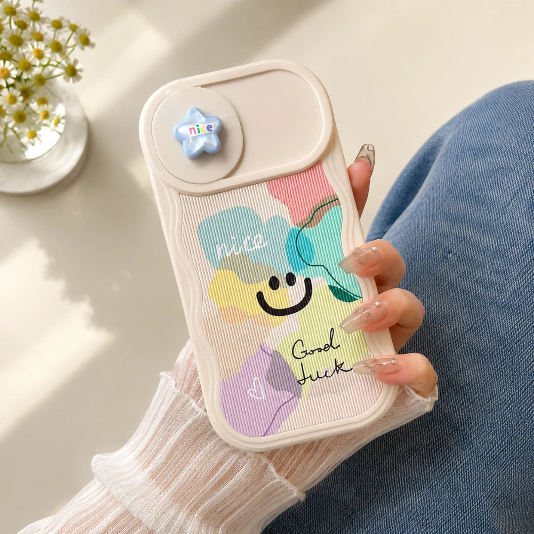 Josh & Tate - CloudPop™ Sliding Lens iPhone Case - A hand holding a pastel-colored phone case with a rainbow cloud design, smiley face, and the words 'nice' and 'good luck'. The case has a small blue star-shaped charm on the back.