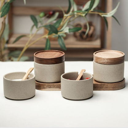 Set of ceramic containers with wooden lids on a white surface, with a plant in the background.