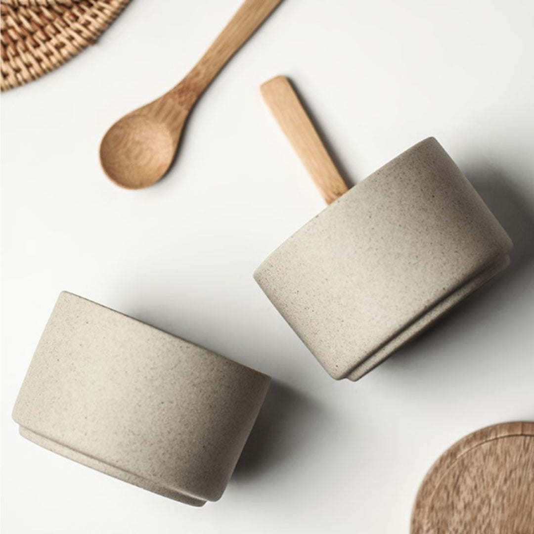 Two beige ceramic cups with wooden handles on a white surface, surrounded by wooden utensils.