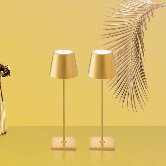 Two gold table lamps on a yellow surface with a palm leaf in the background