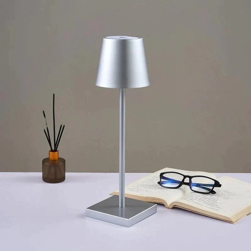 Silver table lamp on a surface with a book, glasses, and a vase in the background.