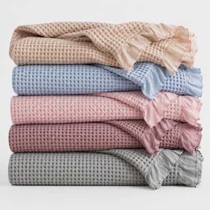 Josh & Tate - CozyCradle™ Ruffle-Edge Cotton Bed Blanket - Stack of folded textured blankets in various colors on a light background