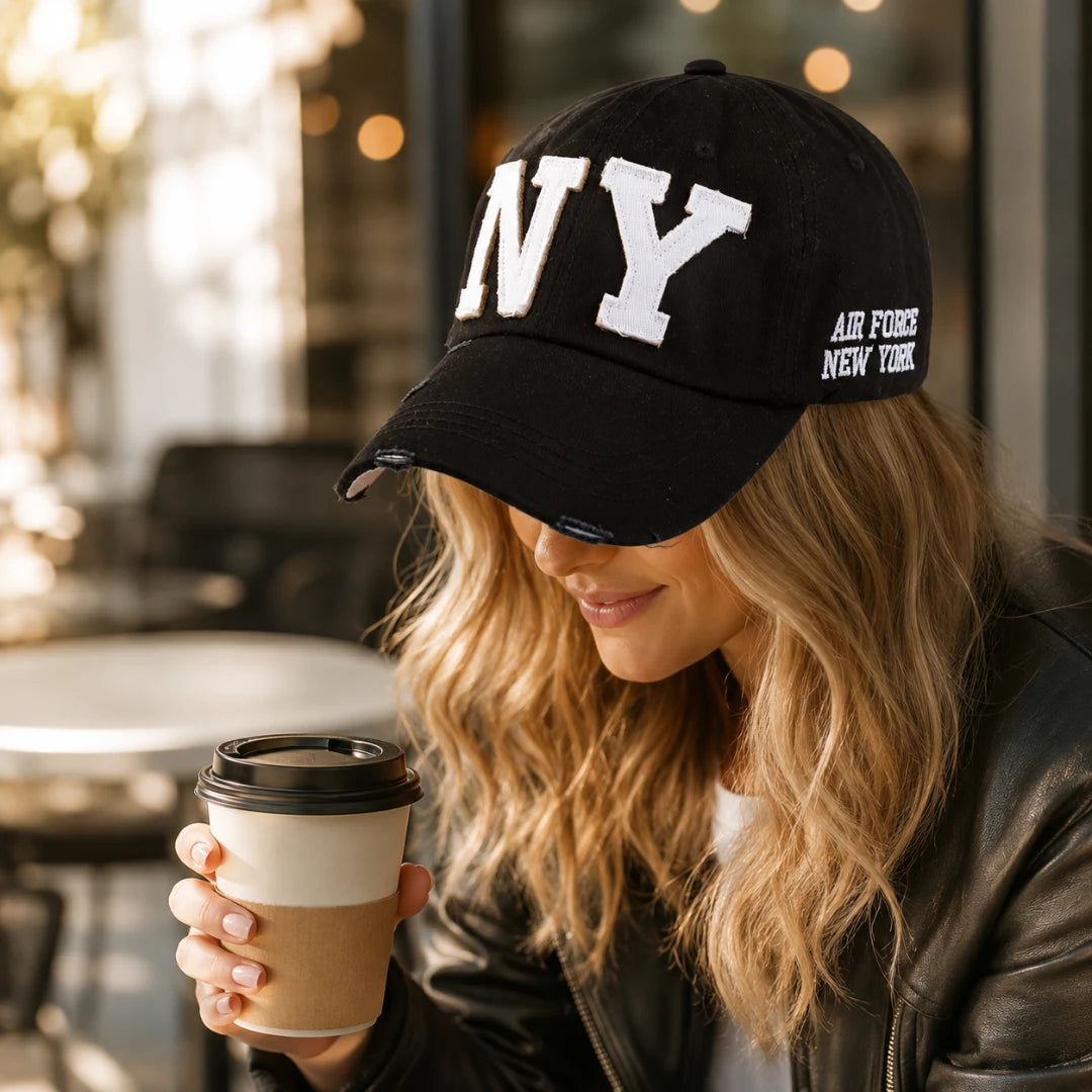 Josh & Tate - CraftWorn™ Distressed NY Cap - A woman wearing a black baseball cap with white embroidered 'NY' lettering on the front and 'AIR FORCE NEW YORK' on the side, holding a paper coffee cup in a cafe setting.