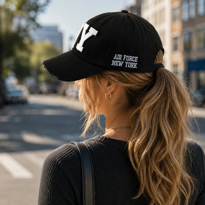 Josh & Tate -CraftWorn™ Distressed NY Cap - A woman seen from behind wearing a black baseball cap with white embroidered lettering that reads 'AIR FORCE NEW YORK' and a large white 'Y' on the front. The background shows a blurred city street scene.