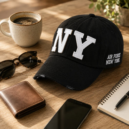 Josh & Tate - CraftWorn™ Distressed NY Cap - A black baseball cap with large white embroidered 'NY' lettering on the front and 'AIR FORCE NEW YORK' on the side, styled on a wooden desk with a coffee mug, sunglasses, wallet, phone, and notebook.
