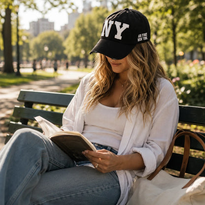Josh & Tate - CraftWorn™ Distressed NY Cap - A woman sits on a park bench reading a book, wearing a black baseball cap with white 'NY' lettering and 'AIR FORCE NEW YORK' text, a white shirt, and blue jeans.