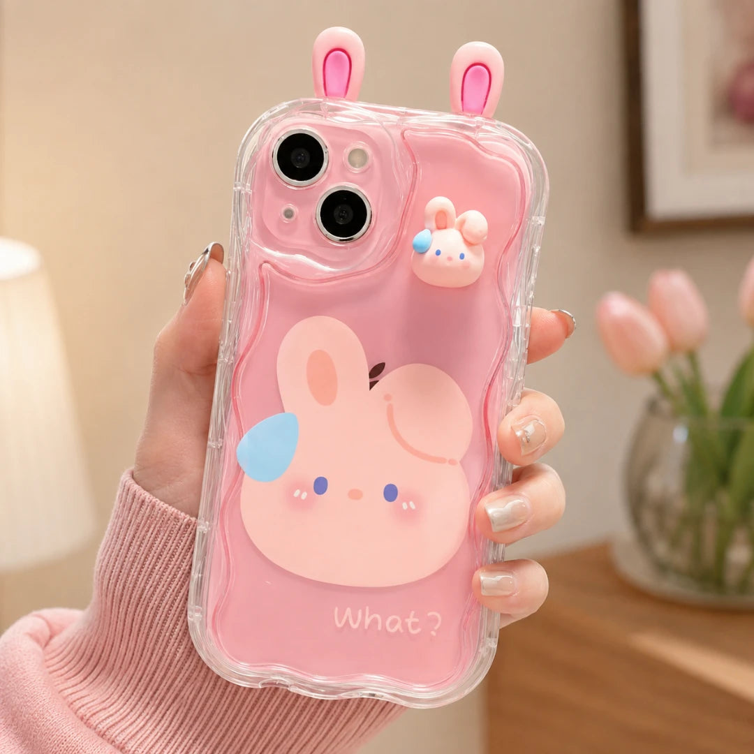 Josh & Tate - CuteSilicone™ iPhone Case - A hand holding a clear phone case decorated with a pink cartoon bunny face, pink bunny ears on top, and a small 3D bunny charm, set against a softly blurred indoor background with pink tulips.