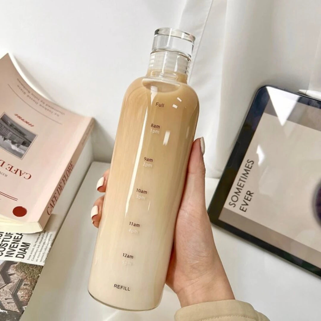Josh & Tate - Daily Water Tracker Bottle - Hand holding a beige water bottle with measurement markings on a surface with books and a tablet.