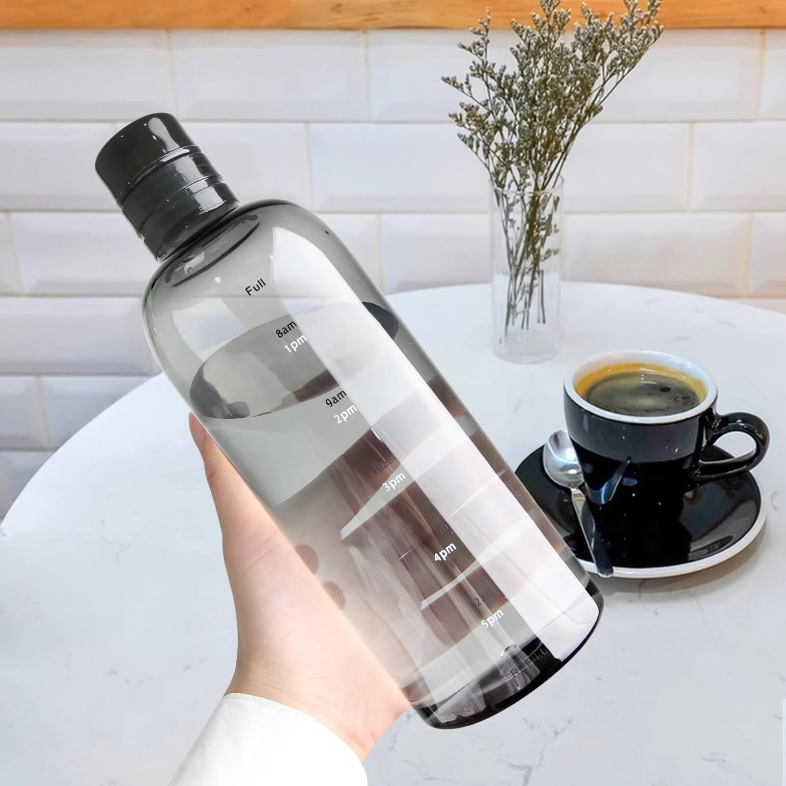 Josh & Tate - Daily Water Tracker Bottle - Clear water bottle with black cap held by a hand, next to a cup of coffee on a table.