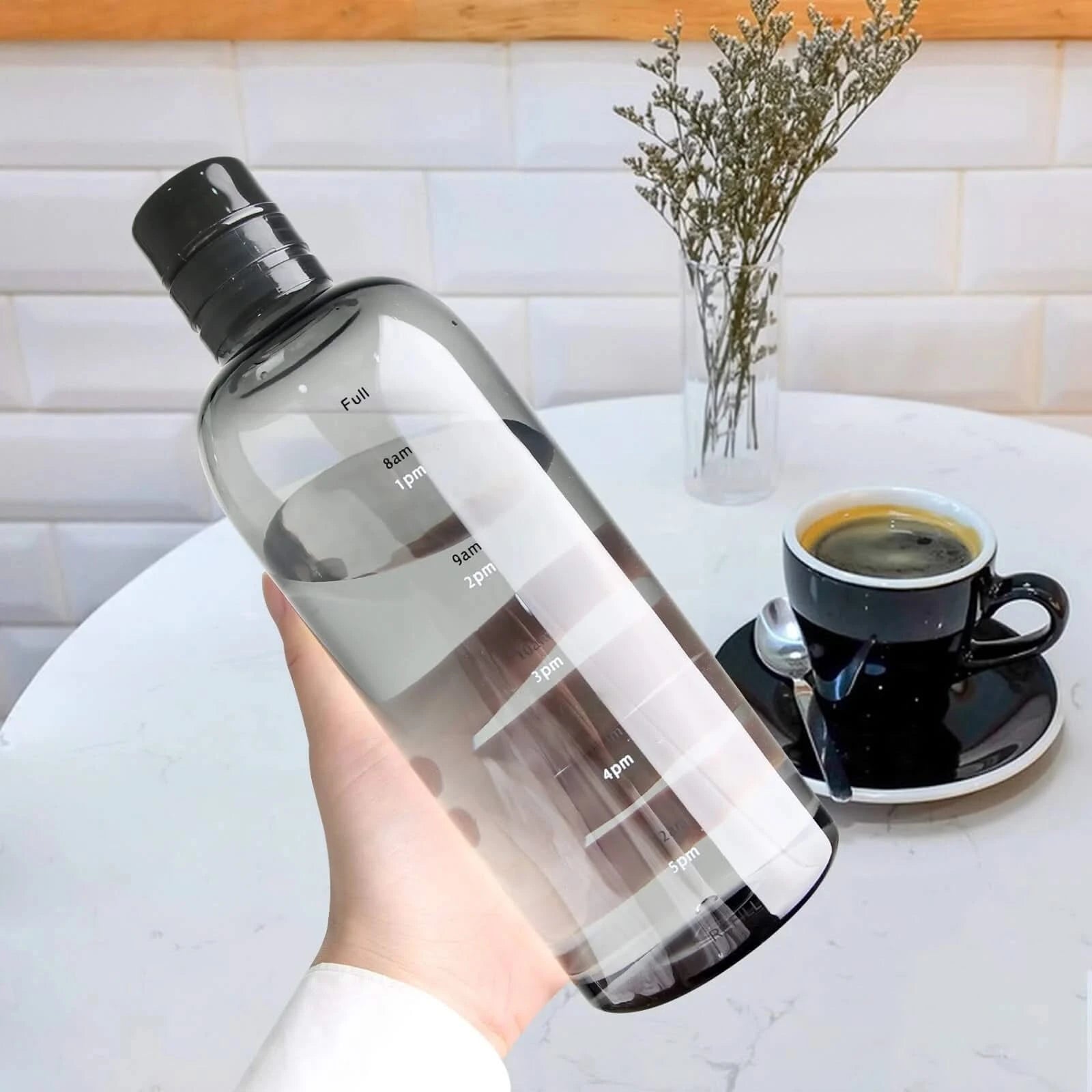 Josh & Tate - Daily Water Tracker Bottle - Clear water bottle with black cap held by a hand, next to a cup of coffee on a table.