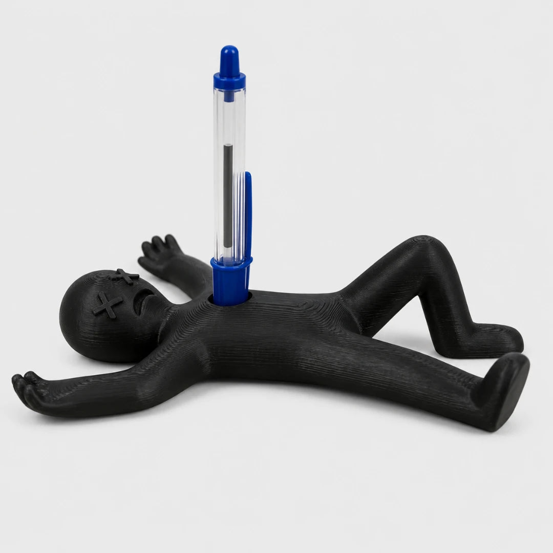 Josh & Tate - DeadDrop™ Desk Pen Stand - A novelty desk accessory featuring a black voodoo doll figure lying on its back with a blue and clear pen inserted into its chest, set against a light gray background.