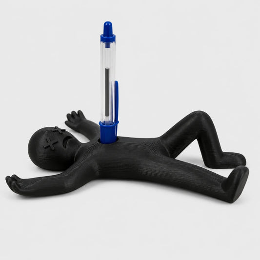 Josh & Tate - DeadDrop™ Desk Pen Stand - A novelty desk accessory featuring a black voodoo doll figure lying on its back with a blue and clear pen inserted into its chest, set against a light gray background.