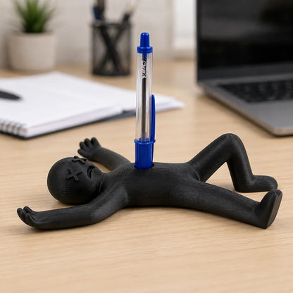 Josh & Tate - DeadDrop™ Desk Pen Stand - A black plastic desk organizer shaped like a person lying on their back, holding a blue and clear pen vertically in the center of the back. The pen has 'LOTUS' printed on it.