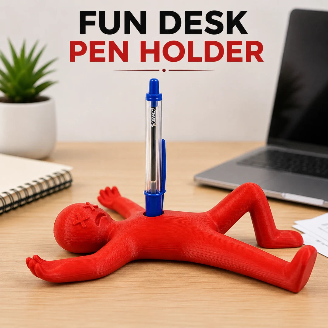 Josh & Tate DeadDrop™ Desk Pen Stand - A red novelty desk pen holder shaped like a lying person, with a blue pen inserted into the torso, shown on a wooden desk with a laptop, notebook, and plant in the background. Large text at the top reads 'FUN DESK PEN HOLDER'.