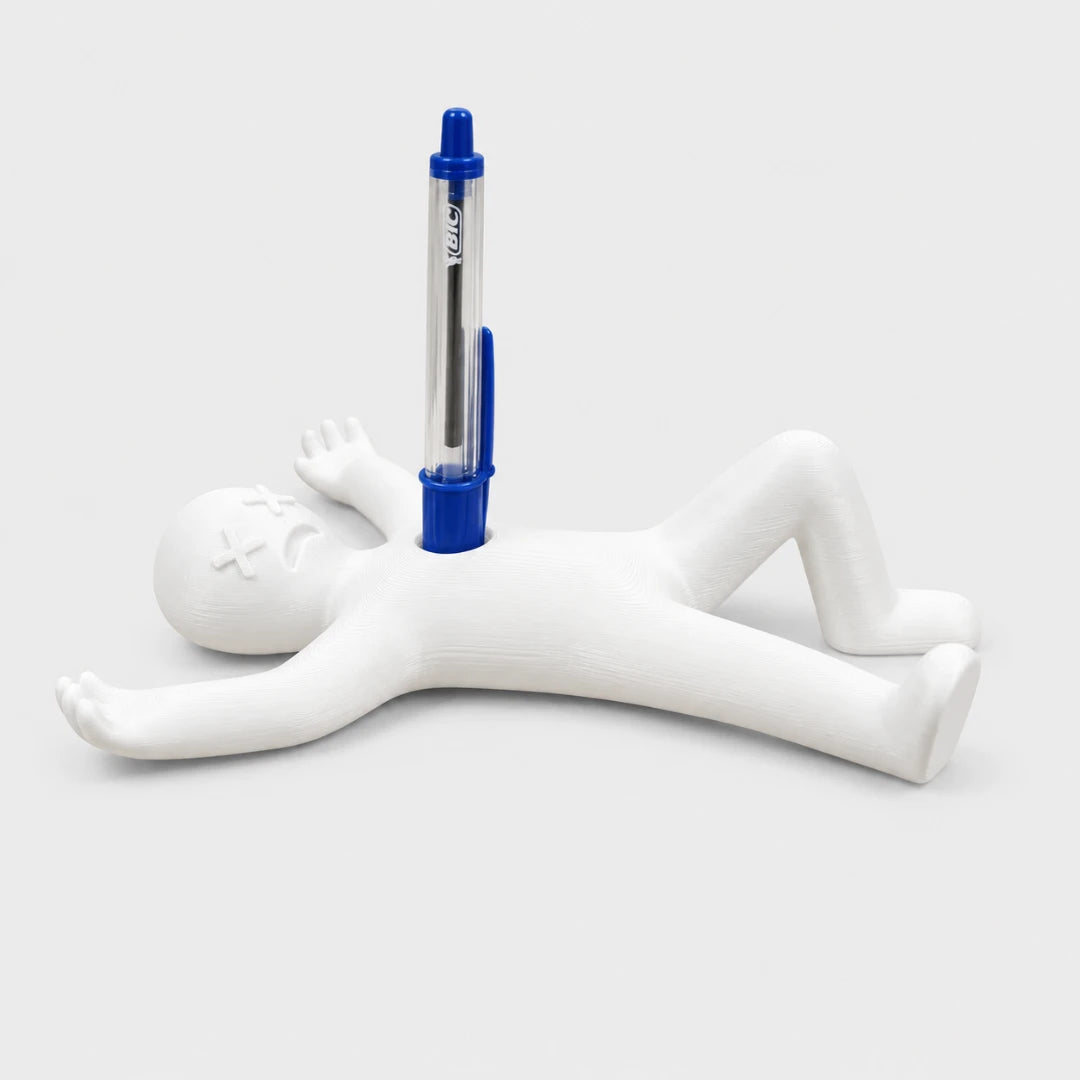 Josh & Tate - DeadDrop™ Desk Pen Stand - A white figurine of a person lying on their back with their arms and legs extended, holding a blue and silver pen vertically between their chest and head. The pen features the brand name 'KAWS' printed on its barrel.