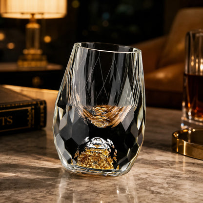 Josh & Tate - DiamondCrystal™ Shot Glass - A clear faceted crystal wine glass with a gold glittery base, shown on a marble tabletop in a warmly lit interior setting with a lamp and books in the background.