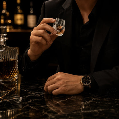 Josh & Tate - DiamondCrystal™ Shot Glass - A man in a black suit jacket and shirt holds a small clear glass tumbler with amber liquid, seated at a dark marble bar counter. A whiskey decanter sits to the left, and he wears a gold-tone wristwatch.